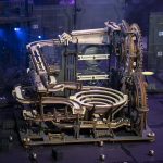Komplexe mechanische Schwerkraftmaschine aus Holz und Metall mit Zahnrädern und Spiralen, die auf einer dunklen Bühne präsentiert wird.