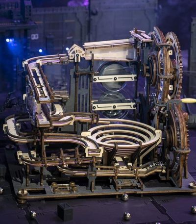 Komplexe mechanische Schwerkraftmaschine aus Holz und Metall mit Zahnrädern und Spiralen, die auf einer dunklen Bühne präsentiert wird.