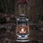 Eine alte Metalllaterne mit brennender Kerze, gehalten über einem Fluss in einem Wald, schafft eine warme und nostalgische Atmosphäre.