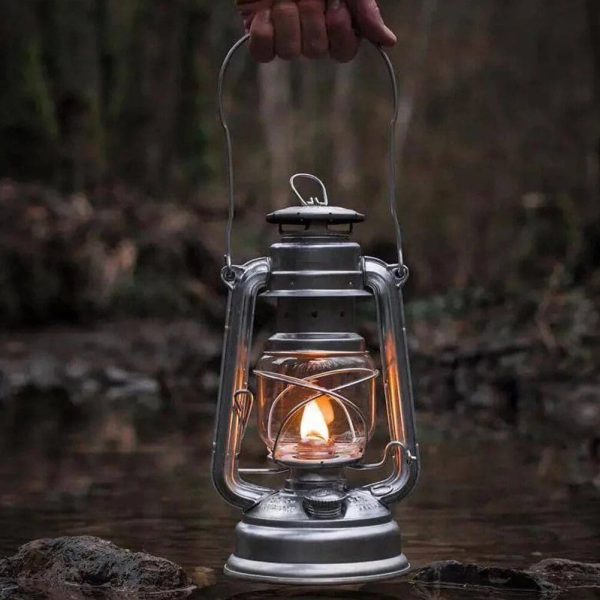 Eine alte Metalllaterne mit brennender Kerze, gehalten über einem Fluss in einem Wald, schafft eine warme und nostalgische Atmosphäre.