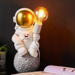 Niedliche Raumfahrer-Figur mit goldenem Helm, die eine Glühbirne hält, sitzt auf einer Mond-Statue. Perfekt als wissenschaftliches Dekorationsstück oder Geschenk für Science-Fans.