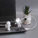 Zwei kleine Astronauten-Spielzeugfiguren auf einem Laptop, verbunden durch ein flexibles Kabel, mit einer Pflanze im Glas im Hintergrund.