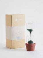 Modernes Sanduhr-Experiment mit schwarzem Sand in einem transparenten Glas, das auf einem roten Blumentopf steht, neben einer Verpackung.