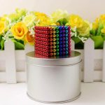 Bunte Magnetperlen in einem Würfelmuster, dekorativ auf einem silbernen Metall-Behälter, vor einem weißen Zaun mit gelben und weißen Blumen im Hintergrund.