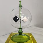 Dekoratives wissenschaftliches Objekt in einer transparenten Glaskugel mit grünen Akzenten, auf einem gelben Sockel präsentiert.