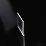 Eine dünne, transparente Glasplatte steht auf einer schwarzen Oberfläche, reflektiert leicht und hebt sich klar vom dunklen Hintergrund ab.