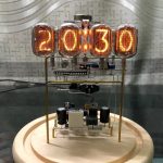 Vintage-Uhr mit digitaler Anzeige, die die Zeit 20:13:00 zeigt, auf einem runden Holzsockel, im Hintergrund eine graue Wand mit geometrischem Muster.