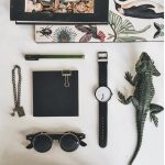 Verschiedene Büro- und Dekorationsgegenstände, darunter eine Armbanduhr, eine Brille, ein Notizbuch, ein Krokodil-Figur und ein Schmuckstück, auf einem weißen Tisch.