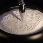 Nahaufnahme eines wissenschaftlichen Experiments mit Flüssigkeit, bei dem ein Metallstab in eine weiße, cremige Substanz eingetaucht wird, um Muster zu erzeugen.