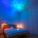 Sternenhimmel-Projektor an der Decke eines Schlafzimmers, der einen leuchtenden Sternenhimmel mit blauen und grünen Lichtern projiziert, schafft eine entspannte Atmosphäre.