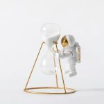 Kleine Astronautenfigur neben einer Sanduhr mit weißem Sand, auf einem eleganten goldenen Rahmen, symbolisiert Wissenschaft und Raumfahrt.