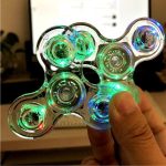 Hand hält einen transparenten, leuchtenden Fidget Spinner mit grünen und blauen LED-Lichtern, vor einem Computerbildschirm im Hintergrund.