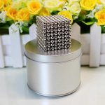 Ein 3D-Magnetwürfel aus kleinen Stahlkugeln, die zu einem glänzenden, geometrischen Kunstwerk verbunden sind, auf einer silbernen runden Box mit einer Blumenrückwand.