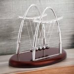 Klassischer Newtons Cradle mit sechs Metallkugeln auf Holzbasis, symbolisiert physikalische Gesetze und Bewegung.