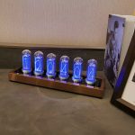 Holographische Zahlenanzeige mit Nixie-Röhren, die die Zahlen 1, 2, 0, 4, 0, 4 in blauer Beleuchtung zeigen, auf einem Holzsockel neben einem Buch und einem Rahmen.