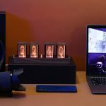 Moderne Nixie-Röhren als wissenschaftliches Dekor auf einem schwarzen Sockel, mit Laptop und Smartphone auf einem Schreibtisch.