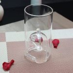 Transparentes Glas mit einer Schnecke im Inneren, umgeben von roten Dekorationselementen, auf einer gestreiften Unterlage.
