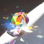 Funkelnder Kristall, der Licht in Regenbogenfarben bricht, auf einer weißen Oberfläche mit Schatten und Lichtreflexen.