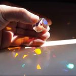 Hand hält einen klaren Kristall, der Licht bricht und bunte Reflexionen auf die Oberfläche projiziert, in einer dunklen Umgebung.