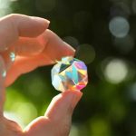 Hand hält einen prismatischen Kristall, der im Sonnenlicht schillert, vor einem unscharfen grünen Hintergrund.
