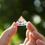 Hand hält einen transparenten Kristallprisma, das im Sonnenlicht schimmert und Regenbogenfarben reflektiert, vor einem unscharfen grünen Hintergrund.