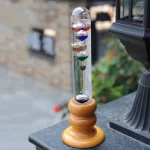 Dekoratives wissenschaftliches Pendel mit bunten Kugeln in einem transparenten Glaszylinder auf Holzsockel, ideal für Bildung und Raumdekoration.