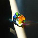 Ein prismatischer Kristall, der Licht in Regenbogenfarben bricht, auf einer dunklen Oberfläche mit Schatten und Lichtreflexionen.
