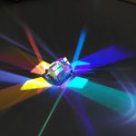 Ein kleines, transparentes Glas, das Licht in bunten Farben bricht und reflektiert, erzeugt ein faszinierendes Farbenspiel auf dunklem Hintergrund.