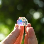 Hand hält einen kleinen, prismatischen Kristall, der im Sonnenlicht schimmert und Regenbogenfarben reflektiert, vor einem unscharfen grünen Hintergrund.