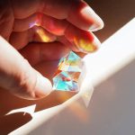 Hand hält einen transparenten Kristall, der Licht in Regenbogenfarben bricht, auf einem weißen Hintergrund.