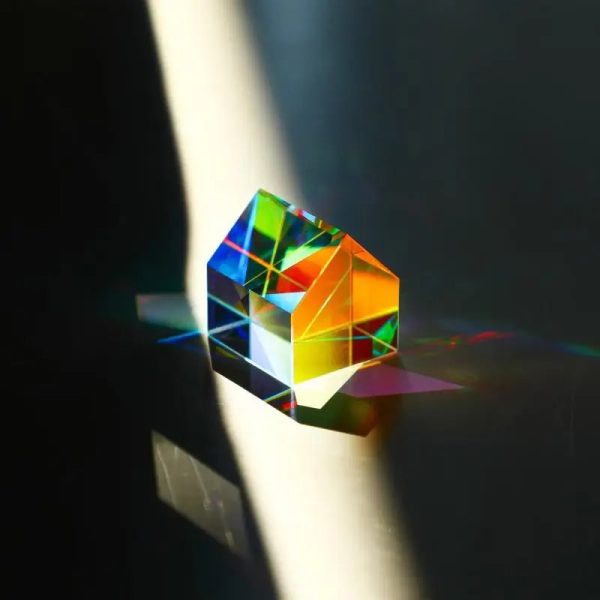 Ein prismatischer Kristall, der Licht in Regenbogenfarben bricht, auf einer dunklen Oberfläche mit Schatten und Lichtreflexionen.