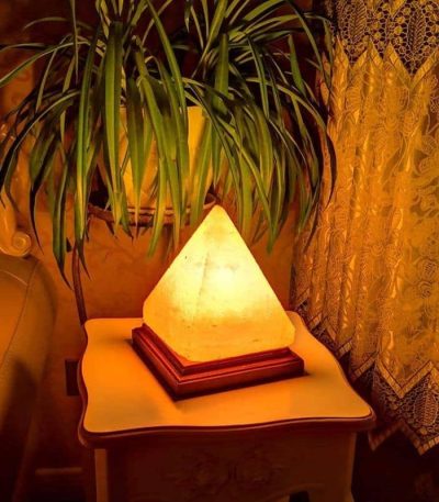 Warm beleuchtete Himalaya-Salzlampe in Pyramidenform auf Nachttisch neben Zimmerpflanze, schafft eine gemütliche Atmosphäre.