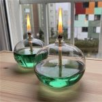 Zwei Kerzen in Glasbehältern mit grüner Flüssigkeit, die Flammen brennen, auf einem Holztisch vor einem Fenster mit Stadtblick.