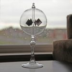 Glasobjekt mit geometrischen Metallteilen, dekorativ auf einem Tisch vor einem Fenster mit Regentropfen, ideal für wissenschaftliche Dekorationen.