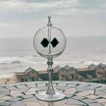 Glasobjekt mit geometrischen schwarzen Elementen, auf einer Mosaikfläche am Strand mit Häusern im Hintergrund, ideal für wissenschaftliche Dekorationen.