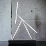 Modernes wissenschaftliches Dekorationsobjekt aus Glasrohren, das an ein chemisches Experiment erinnert, auf einem schwarzen Sockel vor einer Betonwand.