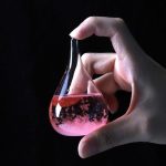 Hand hält ein kleines, pink gefärbtes Reagenzglas mit Flüssigkeit, das wie ein Tropfen geformt ist, vor schwarzem Hintergrund.