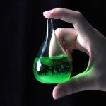 Hand hält ein kleines Glas mit leuchtend grüner Flüssigkeit, das im Dunkeln schimmert.