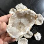 Nahaufnahme eines mineralischen Geode-Kristalls mit weißen, funkelnden Kristallen im Inneren, gehalten in der Hand, mit weiteren Geoden im Hintergrund.
