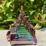 Bunte Kristallskulptur mit schimmernden, metallischen Oberflächen, die in Regenbogenfarben leuchten, vor einem grünen Blätterhintergrund.
