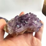 Nahaufnahme eines Amethyst-Kristalls, der auf einer Hand liegt, mit violetten, facettierten Kristallen, die die natürliche Schönheit und Struktur des Minerals zeigen.