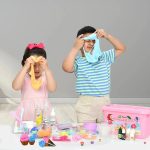 Zwei Kinder spielen mit buntem Schleim auf einem Tisch, die Mädchen haben bunte Kleidung und zeigen Spaß beim Experimentieren.