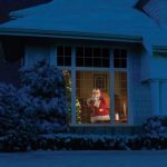 Weihnachtsmann in einem festlich geschmückten Raum, sichtbar durch das Fenster eines Hauses bei Nacht, umgeben von verschneiten Büschen.