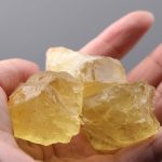 Natürlicher gelber Kristall, gehalten zwischen den Fingern, zeigt eine klare, leuchtende Struktur und eine raue Oberfläche, ideal für wissenschaftliche Dekorationen und Sammlerstücke.
