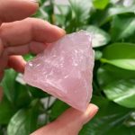 Rohes Rosenquarz-Fragment, handgehalten vor grünen Blättern, natürliche Mineralien, Schmuckstein, Heilstein, Dekoration, Edelstein, Kristall, Naturprodukt, spirituelle Energie.