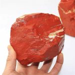 Hand hält einen großen, roten Mineralstein mit weißen und grauen Einschlüssen, ideal als wissenschaftliches Dekorationsstück oder Sammlergegenstand.