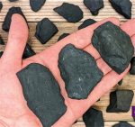 Hand hält mehrere schwarze Gesteinsbrocken, umgeben von weiteren Steinen auf einem Holztisch, ideal für wissenschaftliche oder dekorative Zwecke.