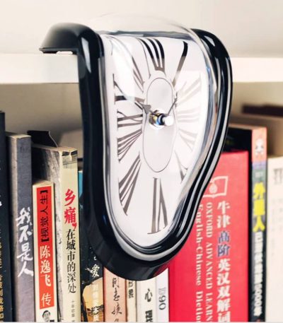 Ein surrealistischer Wecker, der wie geschmolzen wirkt, hängt an einem Bücherregal. Das Design erinnert an Salvador Dalís berühmte Uhren, die die Zeit in einer träumerischen, verzerrten Form darstelle.