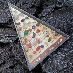 Sammlung verschiedener bunter Steine in einer transparenten Vitrine, die auf einem schwarzen, rauen Gestein liegt.