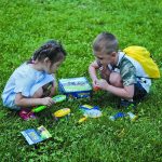 Zwei Kinder spielen mit wissenschaftlichen Entdeckungssets im Grünen, umgeben von bunten Experimentierwerkzeugen und einer Box mit dem Aufdruck "OUTDOOR".